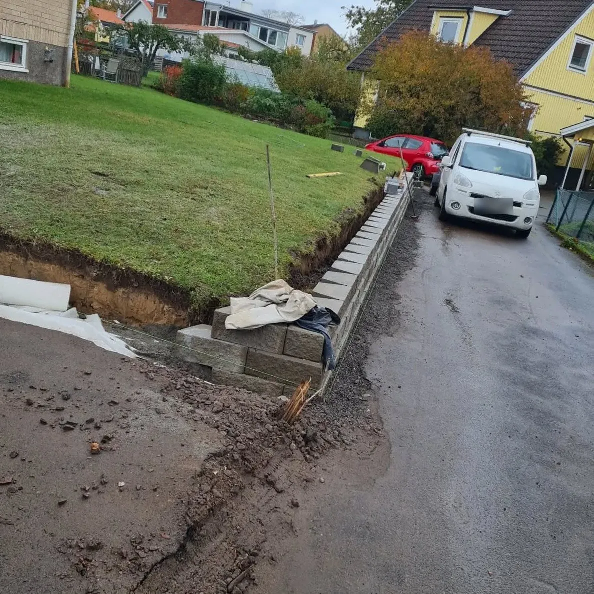 Stödmur av betongblock som byggs längs en uppfart, med schakt och fiberduk mot gräsmattan