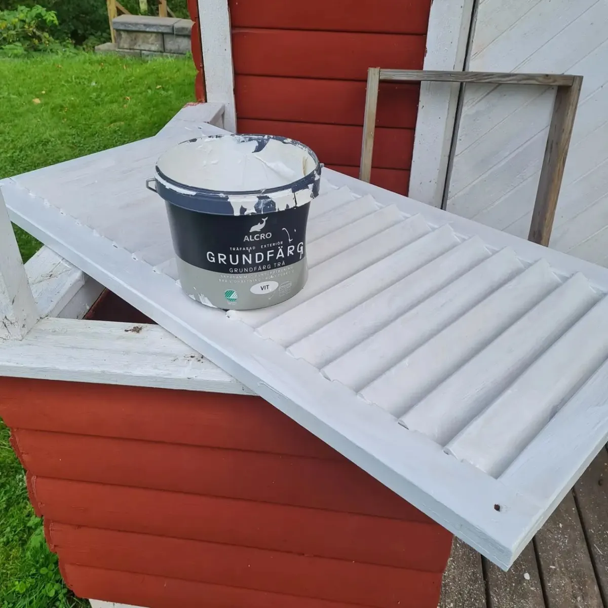 Vit grundmålad fönsterlucka med en färgburk ovanpå, framför en rödmålad bod